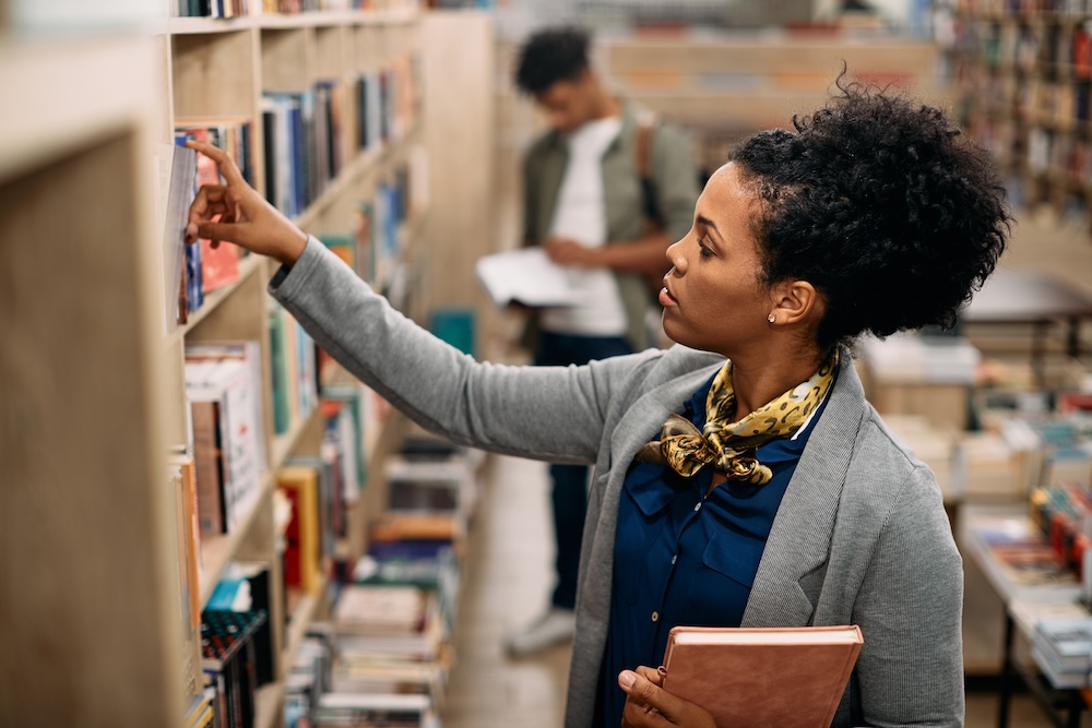 American mid adult student takes book from shelf at university library.