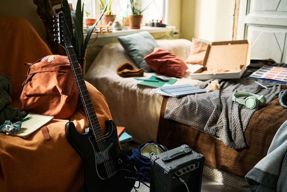 Wide shot of teenager bedroom showing guitar, amplifier, headphones, notebooks, backpack, and pizza box, suggesting creative student lifestyle.
